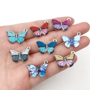 Könnte beinhalten: Sortiment von acht silbernen Schmetterlingsanhängern mit bunten Emailleflügeln. Die Schmetterlinge sind in verschiedenen Farben, darunter Blau, Rosa, Rot und Gelb.