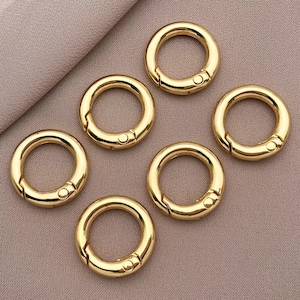 May include: Six gold-colored metal key rings with a spring-loaded clasp. The key rings are arranged in a circle on a light brown surface.