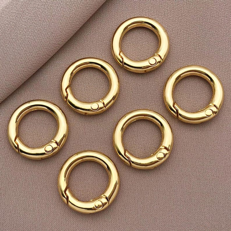 May include: Six gold-colored metal key rings with a spring-loaded clasp. The key rings are arranged in a circle on a light brown surface.