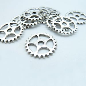 Peut inclure: Un groupe de breloques en forme d'engrenages en métal argenté, chacune avec une taille et une forme différentes. Les engrenages sont disposés de manière aléatoire sur un fond blanc.