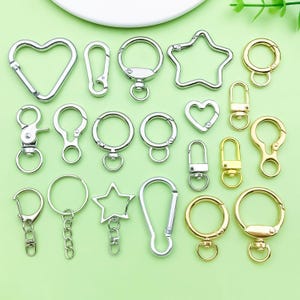 Könnte beinhalten: Eine Auswahl an Metall-Schlüsselanhängern in Silber- und Goldtönen. Die Schlüsselanhänger umfassen Herz-, Stern- und runde Formen sowie Karabiner- und Haken-Stile. Einige haben angebrachte Ringe und Ketten.