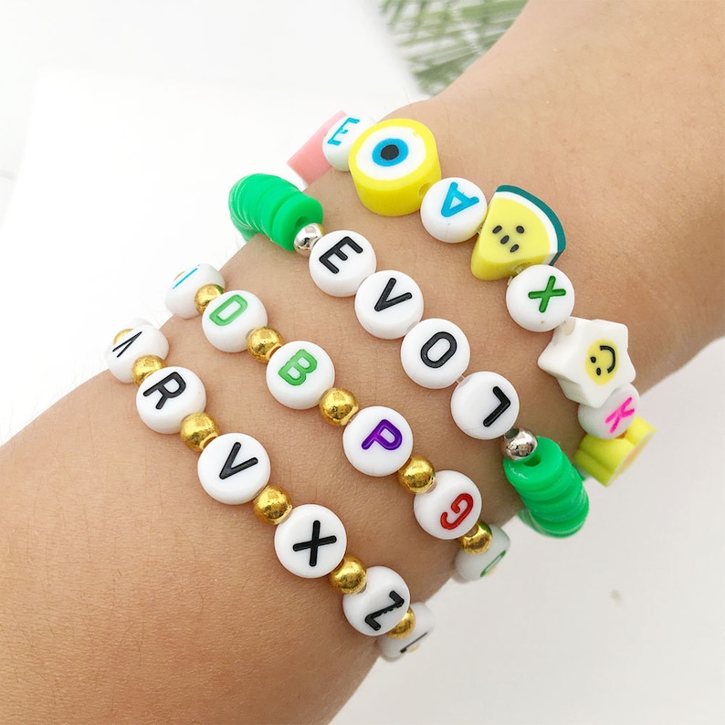 May include: Four colorful bracelets with letter beads, a watermelon bead, an evil eye bead, and a smiley face bead. The bracelets are on a person's wrist.