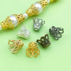 May include: A selection of decorative metal bead caps in gold, silver, and bronze. The caps feature an openwork design and are arranged on a light green surface. A strand of pearls with gold bead caps is visible in the background.