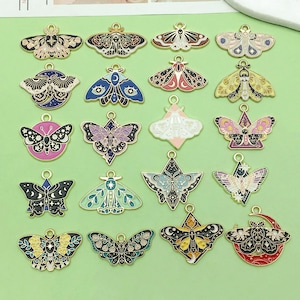 May include: A selection of decorative moth charms in various colours, including gold, blue, pink, and black. Each charm has a detailed design and a small loop for attaching. The charms are displayed on a light green surface, showcasing their intricate patterns.