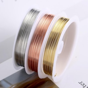 Puede incluir: Tres carretes de alambre, uno plateado, uno de oro rosa y uno dorado, están enrollados alrededor de carretes de plástico blanco. Los carretes están apilados uno encima del otro.