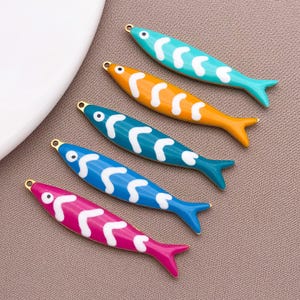 May include: Five colorful fish-shaped charms in turquoise, orange, teal, blue, and pink. Each charm has a white wavy pattern and a gold-colored loop at the top. The charms are arranged on a neutral background.