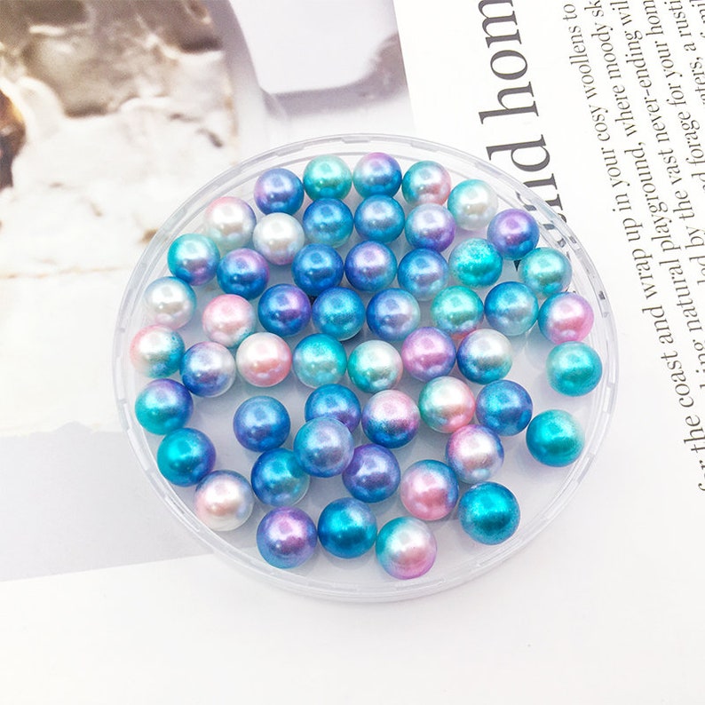May include: A close-up of a clear plastic container filled with iridescent blue, pink, and white pearl beads. The beads are round and have a smooth, shiny finish.