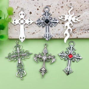 May include: A collection of six silver-colored cross pendants. The crosses vary in design, including ornate filigree, flame-like shapes, and crosses with black or red gemstone accents. Each pendant has a loop for attaching to a chain.