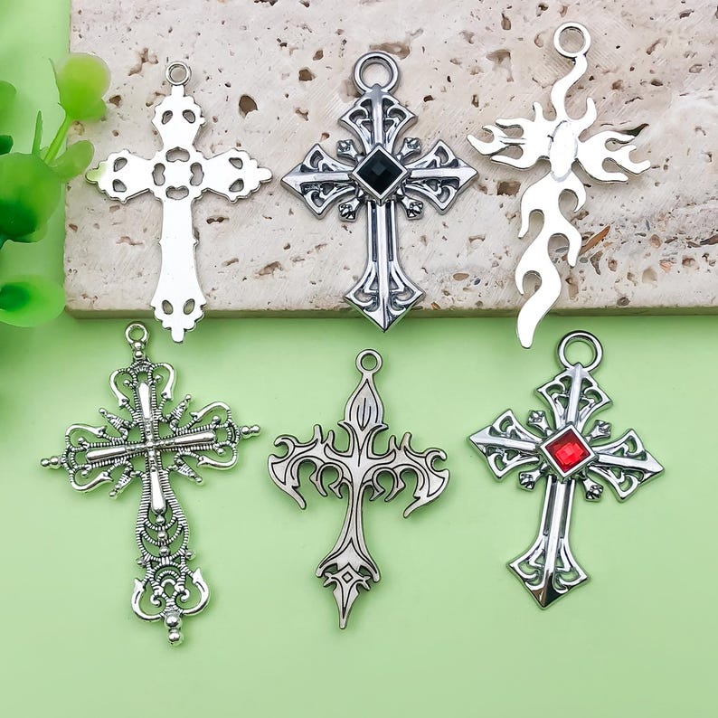 May include: A collection of six silver-colored cross pendants. The crosses vary in design, including ornate filigree, flame-like shapes, and crosses with black or red gemstone accents. Each pendant has a loop for attaching to a chain.