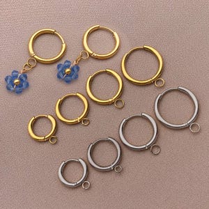 May include: An assortment of gold and silver hoop earrings. Some gold hoops have blue beaded flower charms. The earrings vary in size and have a small loop for attaching to the ear. The earrings are displayed on a light brown surface.