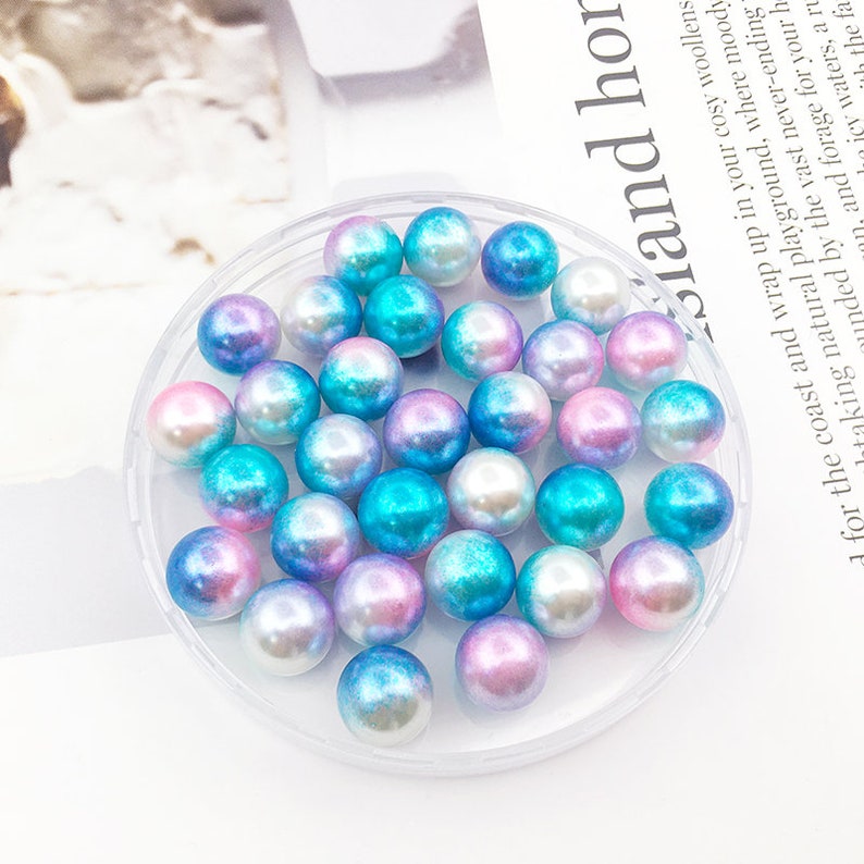 May include: A clear plastic container filled with iridescent blue and pink pearl beads. The beads are round and have a smooth, shiny finish.