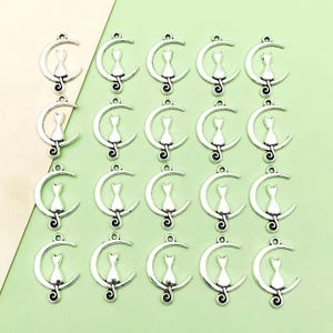 May include: Twenty silver-toned metal charms featuring a cat sitting on a crescent moon. The charms have a hole at the top for attaching to jewellery or other crafts.