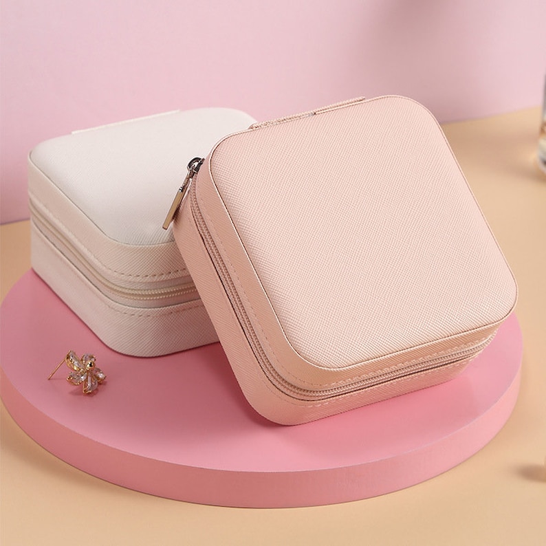 May include: Two square-shaped jewelry boxes with zippers. The box on the left is white and the box on the right is pink.