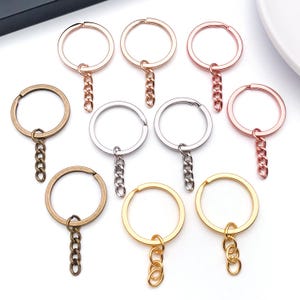 Puede incluir: Una colección de llaveros en varios acabados metálicos, incluyendo dorado, plateado y oro rosa. Cada llavero presenta un anillo circular y una cadena para sujetar llaves. Los llaveros están dispuestos sobre una superficie blanca.