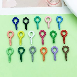 May include: A collection of colorful screw eye hooks in various shades, including blue, black, green, pink, yellow, white, red, gray, orange, purple, and brown. These small, decorative hardware pieces are arranged on a light green surface.
