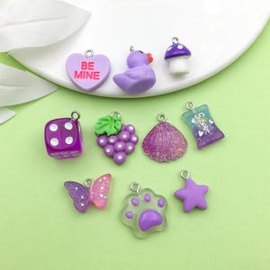Op de afbeelding: Assortiment van paarse en roze harscharms met verschillende ontwerpen, waaronder een hart met de tekst "Be Mine", een tros druiven, een schelp, een dobbelsteen, een vlinder, een pootprint, een ster, een paddenstoel, een eend en een snoepzakje met de tekst "Sweet".