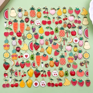 May include: A selection of fruit-shaped charms, such as strawberries, pineapples, cherries, and watermelons. Each charm has a gold-coloured border and a small loop for attaching to jewellery or crafts. The charms are displayed on a light green surface.