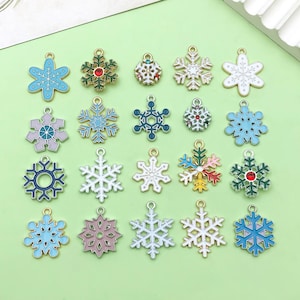 Op de afbeelding: Assortiment van 18 gouden sneeuwvlokcharms met verschillende ontwerpen in gekleurd email. De charms zijn perfect voor het maken van sieraden, scrapbooking en andere knutselprojecten.