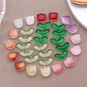 Puede incluir: Una colección de cuentas de vidrio de colores en varias formas. Las cuentas incluyen formas de tulipán translúcidas en tonos de rojo, naranja, rosa y amarillo, y cuentas en forma de mariposa en verde y transparente. Las cuentas están dispuestas sobre una superficie de tono neutro.