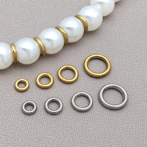 Peut inclure: Divers anneaux métalliques dorés et argentés de différentes tailles, présentés sur une surface texturée. Un collier de perles avec des détails dorés est visible en arrière-plan. Idéal pour la création de bijoux.