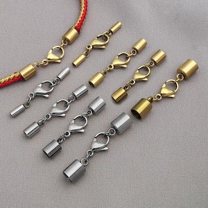 Op de afbeelding: Een assortiment sieraden sluitingen in goud- en zilverkleuren. De sluitingen omvatten karabijnsluitingen en cilindrische eindkappen. Eén sluiting is bevestigd aan een rood en goud gevlochten koord. De sluitingen worden getoond op een neutrale grijze ondergrond.