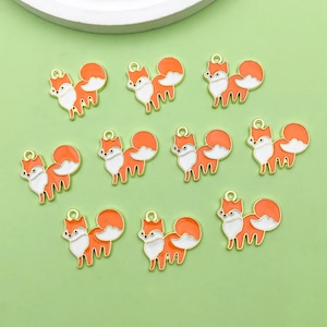 Op de afbeelding: Tien oranje en witte vossencharms met gouden accenten. De vossen staan in verschillende poses, sommige met hun staart omhoog gekruld en andere met hun staart recht uit.