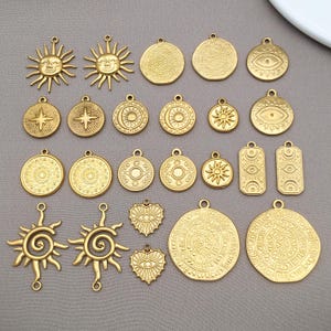 May include: A selection of gold-toned metal charms featuring suns, moons, stars, and eye motifs. The charms display detailed embossed patterns and small loops for attachment. The background is a plain grey colour.