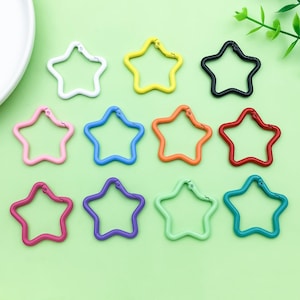 Puede incluir: Una colección de mosquetones en forma de estrella en varios colores: blanco, amarillo, negro, rosa, azul, naranja, rojo, magenta, morado, verde y turquesa. Estos clips metálicos están dispuestos sobre una superficie verde claro.