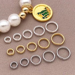 以下が含まれることがあります： さまざまなサイズのシルバーとゴールドカラーの丸カンが、ニュートラルな色の表面に配置されています。カラフルなデザインのゴールドカラーのチャームと、パールとゴールドビーズのブレスレットも表示されています。 ジュエリー作りや工芸に最適です。