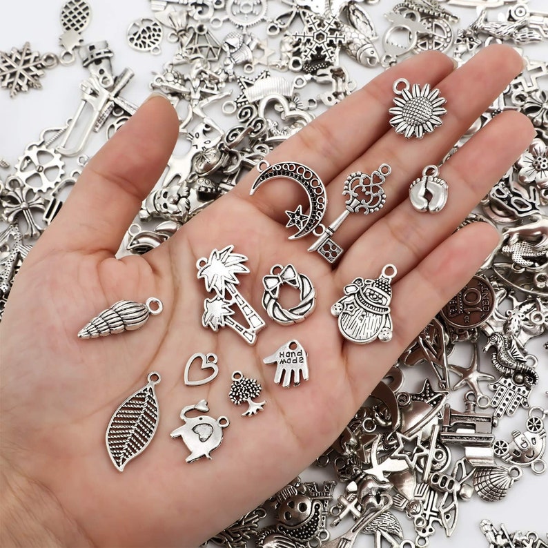Pu&ograve; includere: Una collezione di ciondoli in argento con vari disegni, tra cui una palma, una conchiglia, una luna crescente, un pupazzo di neve, una mano con le parole "Hand Made", una foglia, un cuore, un albero e un elefante.