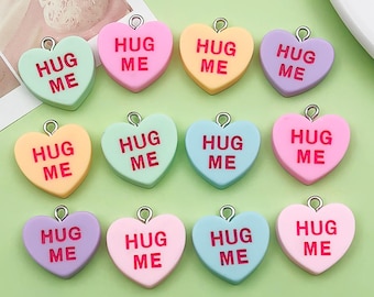 May include: A collection of heart-shaped charms in various pastel colors, each with a silver loop and the words "HUG ME" in bold letters. The charms are arranged on a light green surface.
