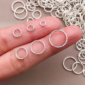Puede incluir: Primer plano de varios anillos pequeños plateados texturizados. Los anillos, de diferentes tamaños, están dispuestos sobre un fondo neutro. Los anillos tienen una textura retorcida en forma de cuerda.