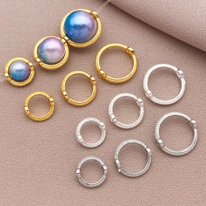 Könnte beinhalten: Ein Satz goldener und silberner Sprungringe, einige mit perlmuttartigem Finish. Die Ringe sind in verschiedenen Größen und auf einer hellbraunen Oberfläche angeordnet.