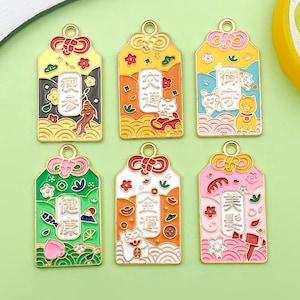 May include: Six colorful metal charms with Japanese text and designs. Each charm has a gold-colored top loop and a rectangular shape with rounded edges. The charms feature various illustrations, including cats, dogs, and flowers, with a variety of colors.