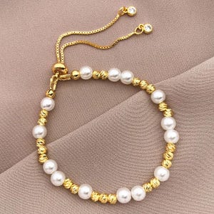 May include: An elegant gold-coloured bracelet with alternating white pearls and textured gold beads. The adjustable design features a delicate chain with two small, clear crystal accents. The bracelet is suitable for formal or casual wear.