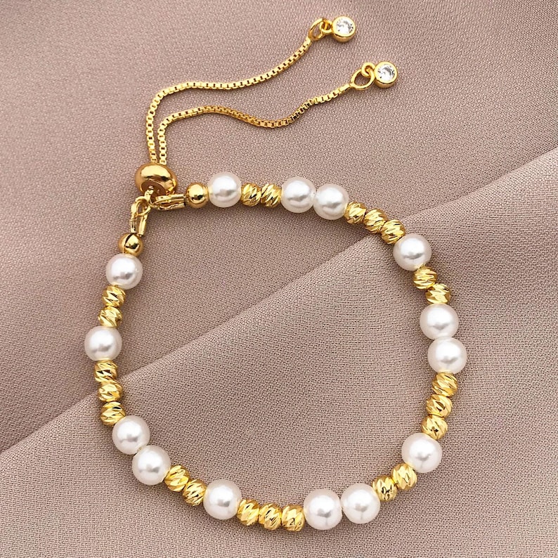 May include: An elegant gold-coloured bracelet with alternating white pearls and textured gold beads. The adjustable design features a delicate chain with two small, clear crystal accents. The bracelet is suitable for formal or casual wear.