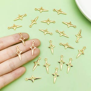 Puede incluir: Una colección de dijes en forma de estrella de metal dorado con centros abiertos. Los dijes están esparcidos sobre una superficie verde claro.