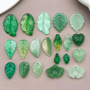 May include: A collection of twenty-two leaf-shaped beads in various shades of green, ranging from translucent pale mint to deep emerald. Each bead features a small hole for stringing, with diverse textures including smooth, matte, and glittery finishes.