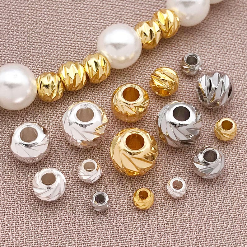 May include: A collection of gold and silver beads in different sizes and styles. Some have a textured, cut appearance, while others are smooth. A strand of large, white pearl beads is also present.
