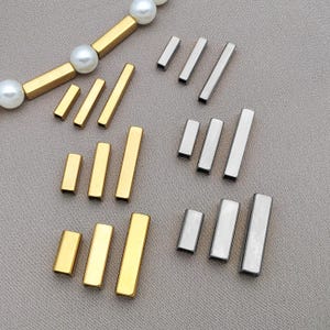 Peut inclure: Divers éléments de bijoux rectangulaires de couleur or et argent. Certains sont disposés par ensembles de trois, d'autres sont dispersés. Un collier avec des barres dorées et des perles blanches est visible.