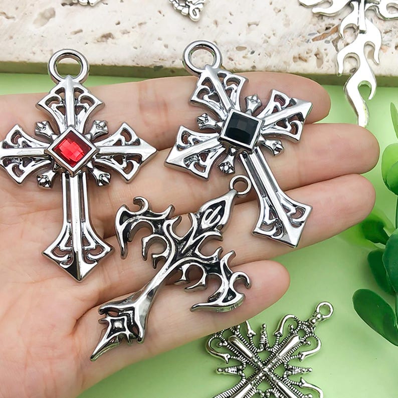 May include: Close-up of several silver-toned cross pendants. Two crosses have a gemstone in the center, one red and one black. The other crosses have intricate, decorative designs. The pendants are held in a hand.