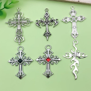 May include: Six silver-toned cross pendants with intricate designs. Some crosses feature decorative embellishments, while others have a gemstone in the center. The crosses vary in style, including gothic and flame designs.