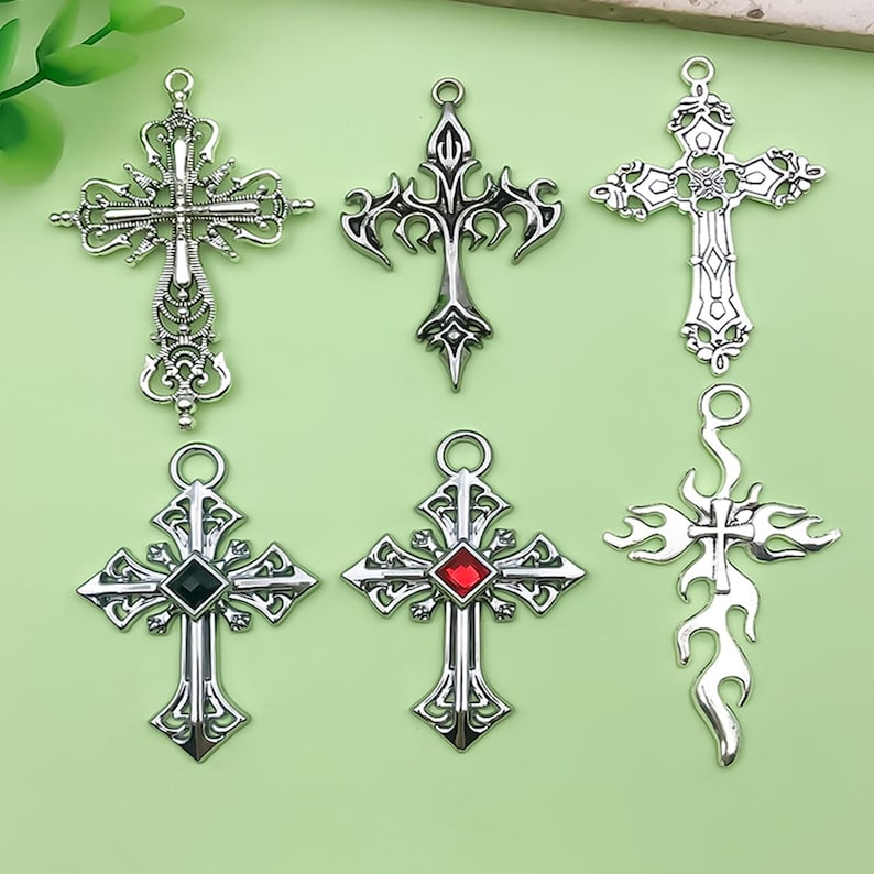 May include: Six silver-toned cross pendants with intricate designs. Some crosses feature decorative embellishments, while others have a gemstone in the center. The crosses vary in style, including gothic and flame designs.