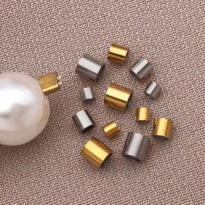 Puede incluir: Un surtido de cuentas tubulares de metal dorado y plateado, junto con una gran perla blanca. Las cuentas varían en tamaño y están dispersas sobre una superficie texturizada de color marrón claro. Estos componentes son probablemente para la fabricación de joyas.