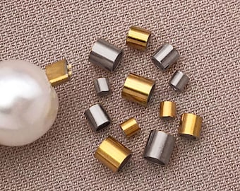May include: An assortment of gold and silver-colored metal tube beads, along with a large white pearl. The beads vary in size and are scattered on a textured, light brown surface. These components are likely for jewelry making.