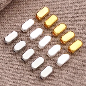 May include: An assortment of metallic beads in gold and silver. The beads are oblong with rounded edges and a hole at each end. They are arranged on a textured, neutral-toned surface.