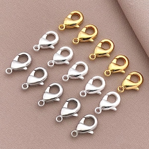 May include: A collection of silver and gold lobster claw clasps, commonly used for jewelry making. The clasps are arranged in a row on a light brown surface.