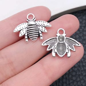 May include: Two silver bee charms with a detailed design. The charms have a loop at the top for attaching to jewelry or other crafts.