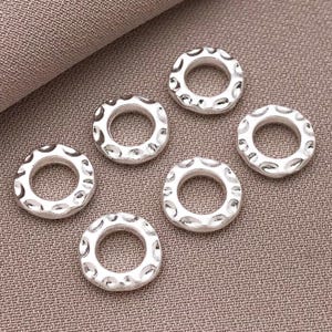 Pu&ograve; includere: Sei perline circolari color argento con un motivo strutturato e puntinato sul bordo esterno. Le perline hanno un foro al centro e sono disposte su una superficie di tessuto dai toni neutri. Ideali per la creazione di gioielli.