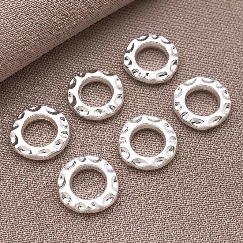 Pu&ograve; includere: Sei perline circolari color argento con un motivo strutturato e puntinato sul bordo esterno. Le perline hanno un foro al centro e sono disposte su una superficie di tessuto dai toni neutri. Ideali per la creazione di gioielli.